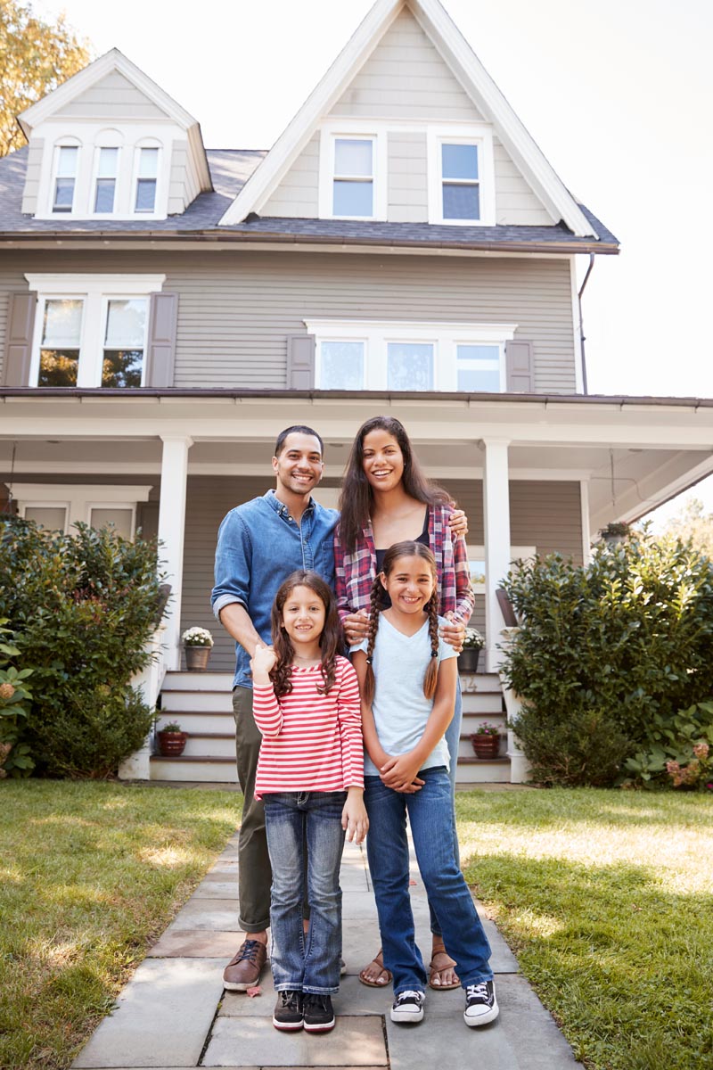 A family of four standing on the front walk of a gray two-story house.